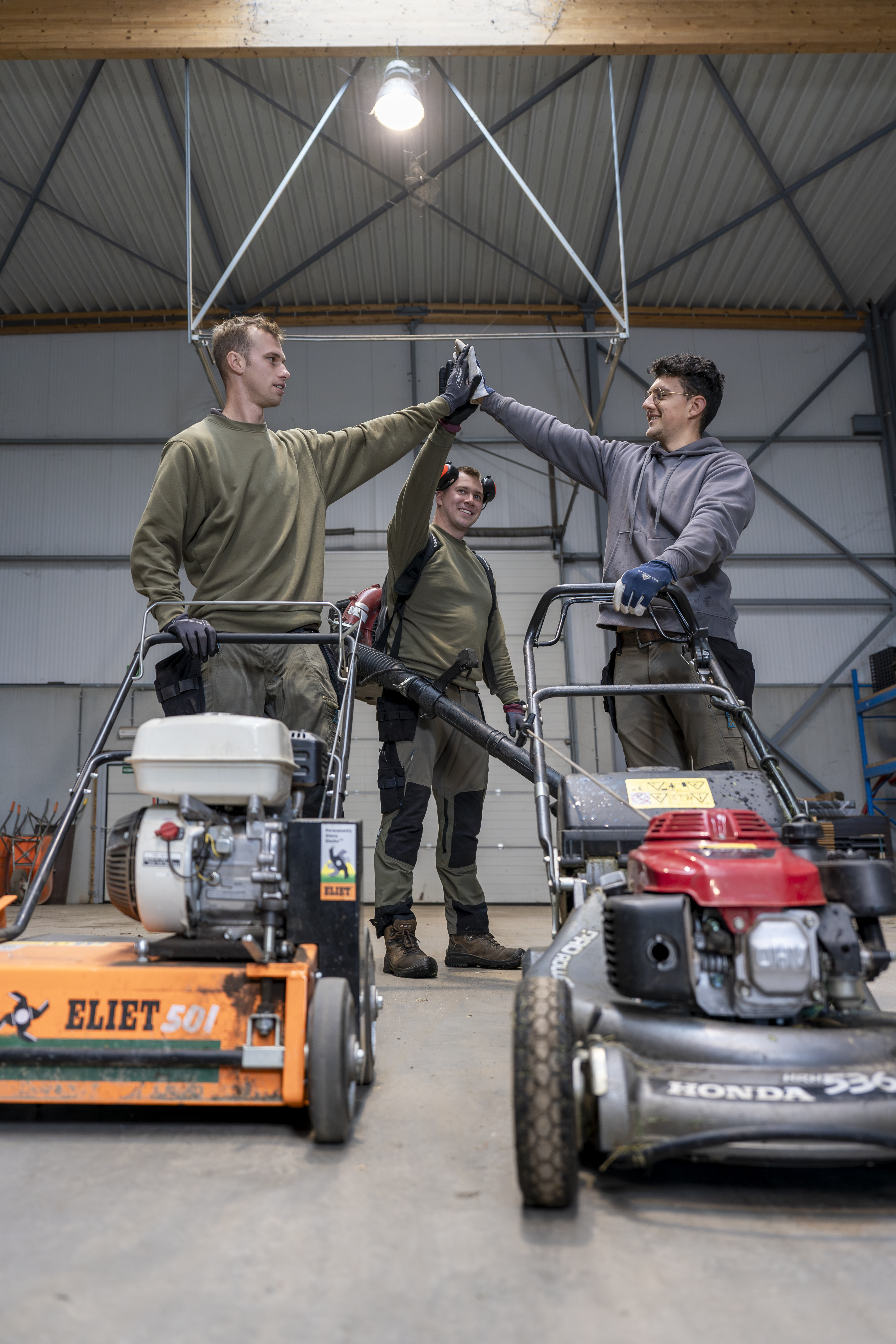 Drie werknemers met de handen in de lucht met grasmaaiers en een bladblazer.